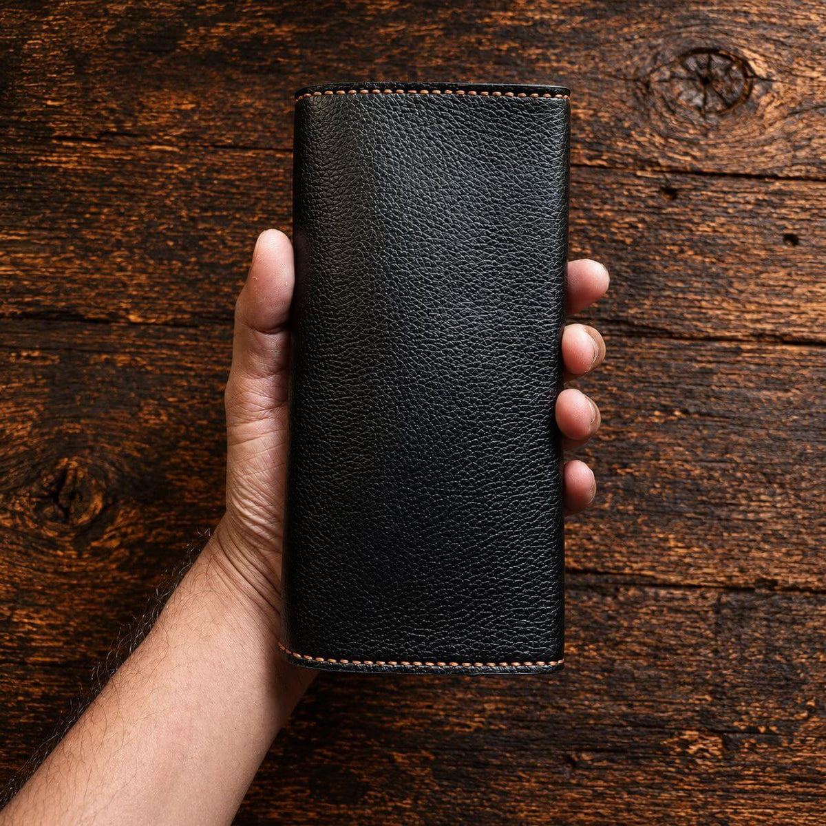 A hand holding a black textured leather wallet or phone case with light tan contrast stitching, set against a rustic dark brown wooden background.