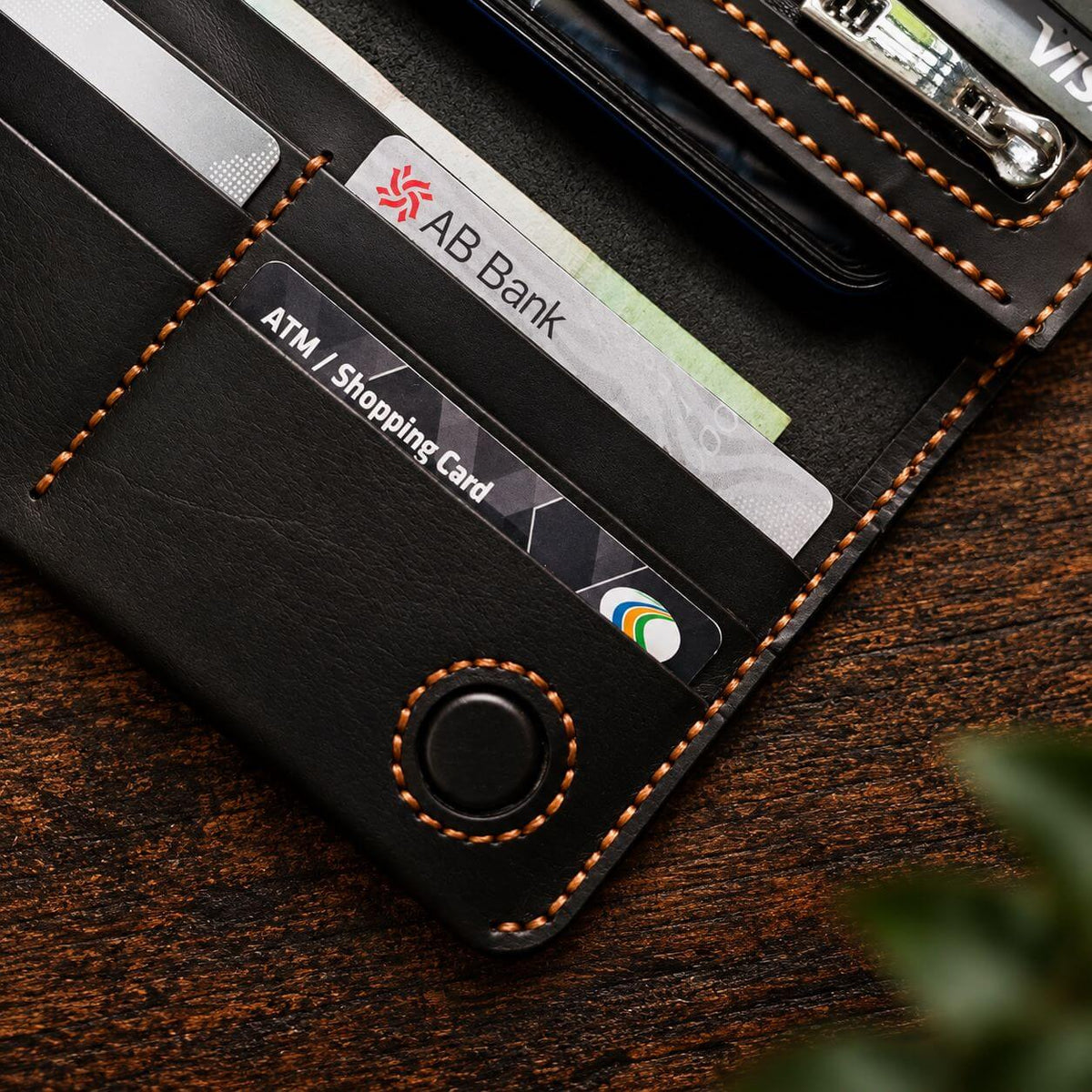 A close-up of a black leather wallet with orange contrast stitching, shown open with cards and a phone partially visible. The wallet is photographed on a dark wood surface with a blurred green plant in the foreground.
