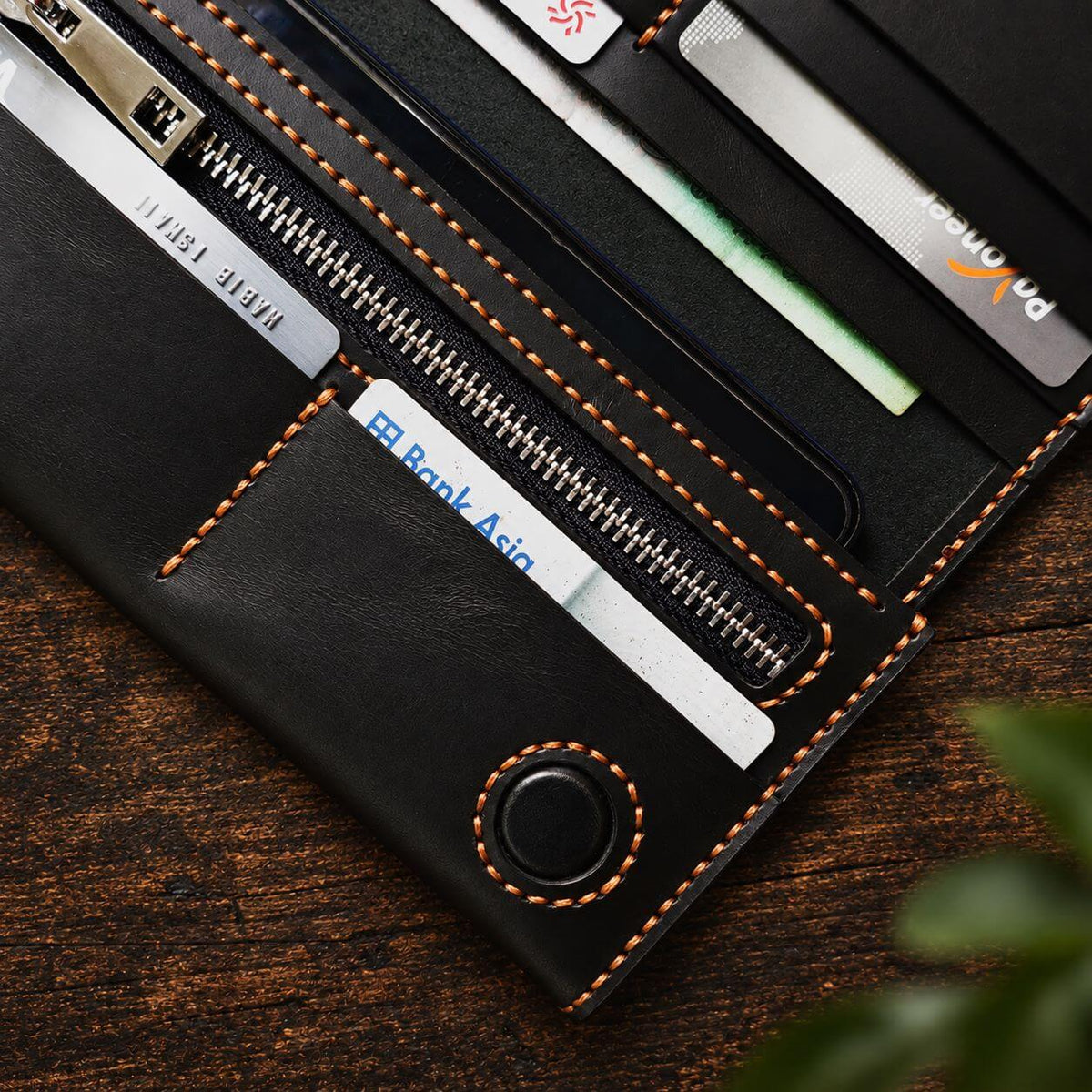 A close-up of an open black leather wallet or card holder with orange contrast stitching, showing card slots, a zip compartment, and inserted cards, placed on a dark wooden surface.
