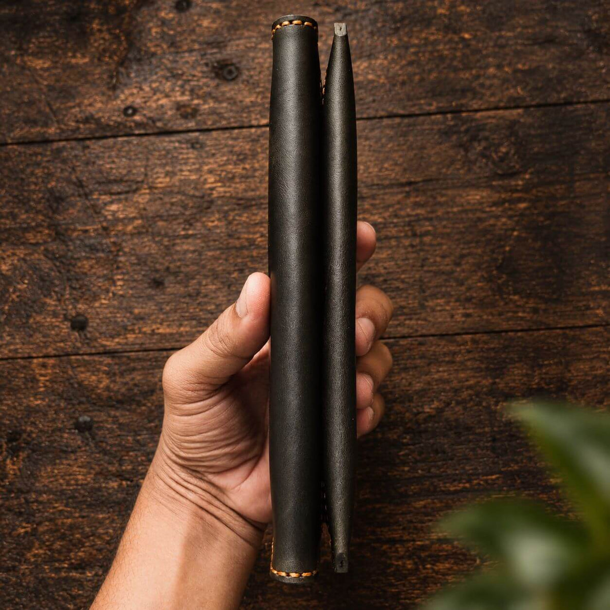 A hand holds a slim black leather wallet or card holder with a matching pen in a vertical orientation, set against a rustic dark wood background.