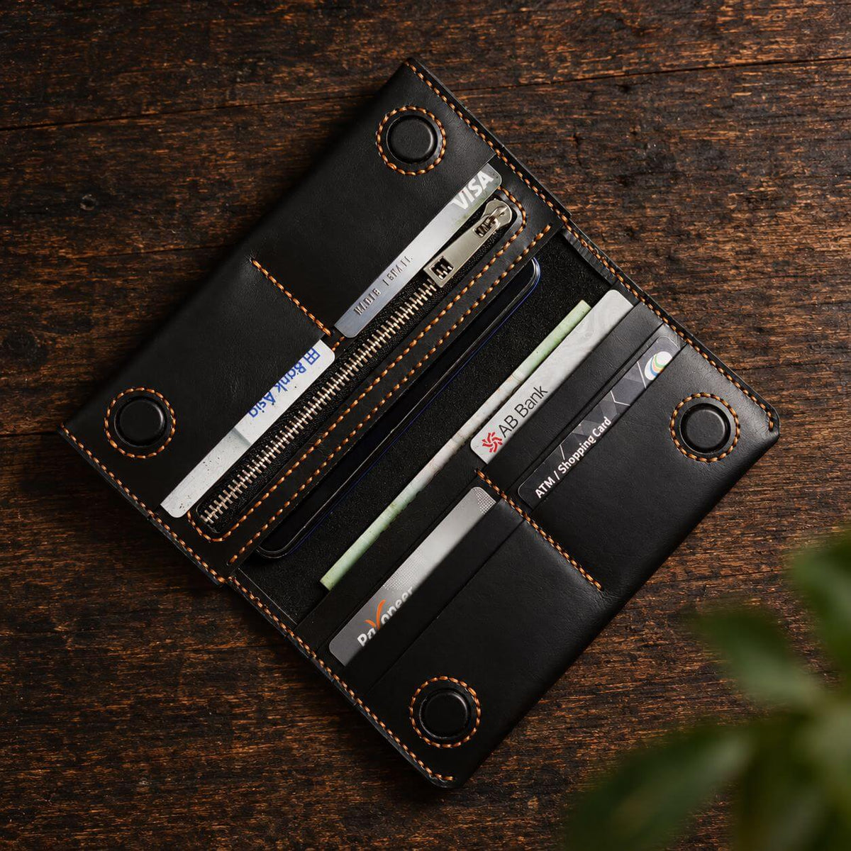 A black leather card holder wallet with orange contrast stitching, shown open on a dark wooden surface. The wallet contains several cards and a phone inside its slots.