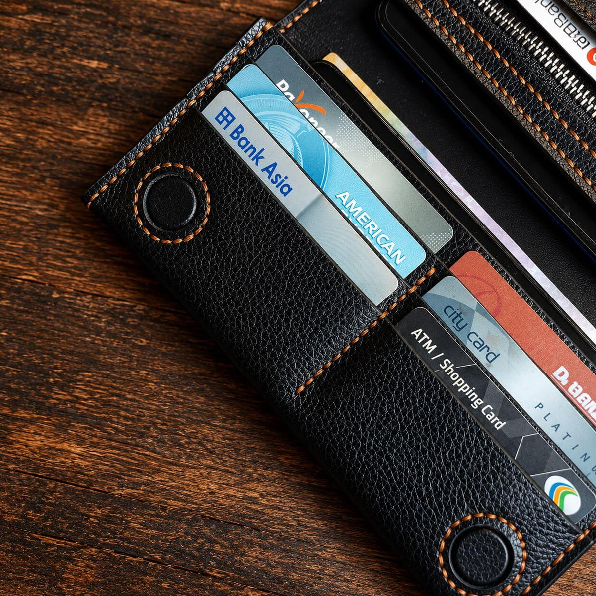 A black pebbled leather bi-fold wallet with orange contrast stitching, shown open on a dark wood surface. Several credit cards are inserted into the card slots, including visible brands like American Express and D.