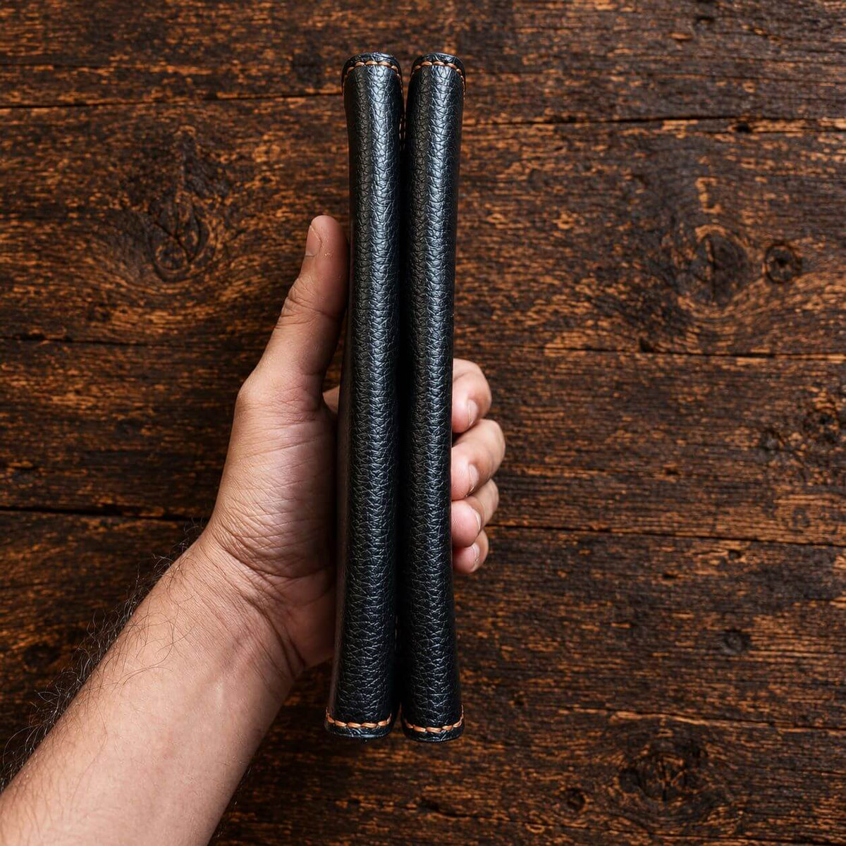 A hand holding two black textured leather cylindrical sleeves or cases against a rustic dark wood plank background.