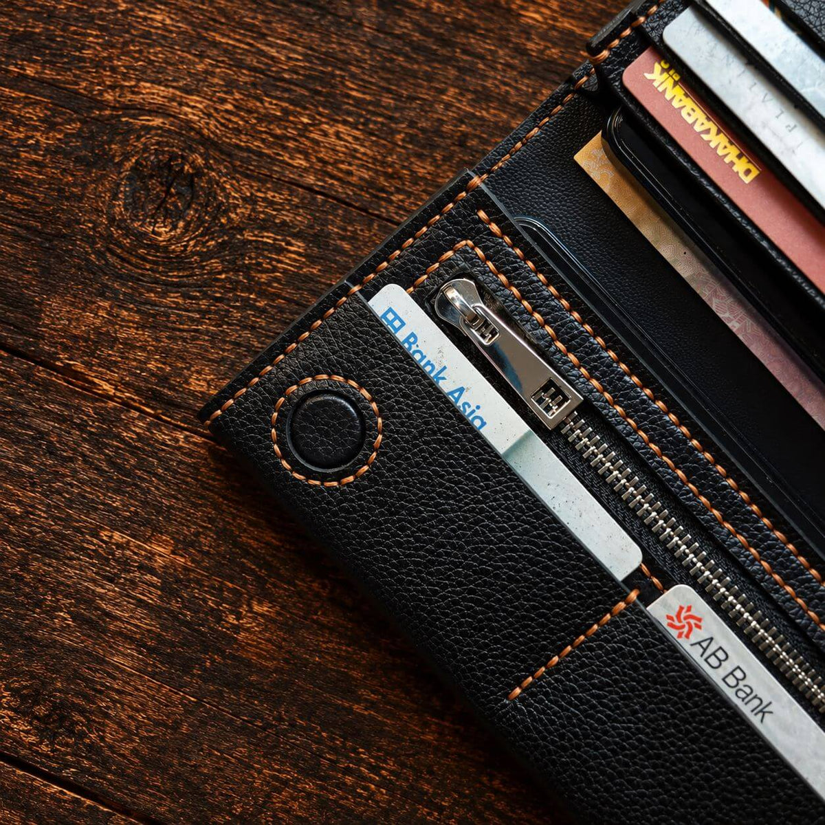 A close-up of a black textured leather wallet with orange contrast stitching, shown open on a dark wood surface. The wallet contains card slots, a zippered compartment, and visible cards, including one labeled AB Bank.