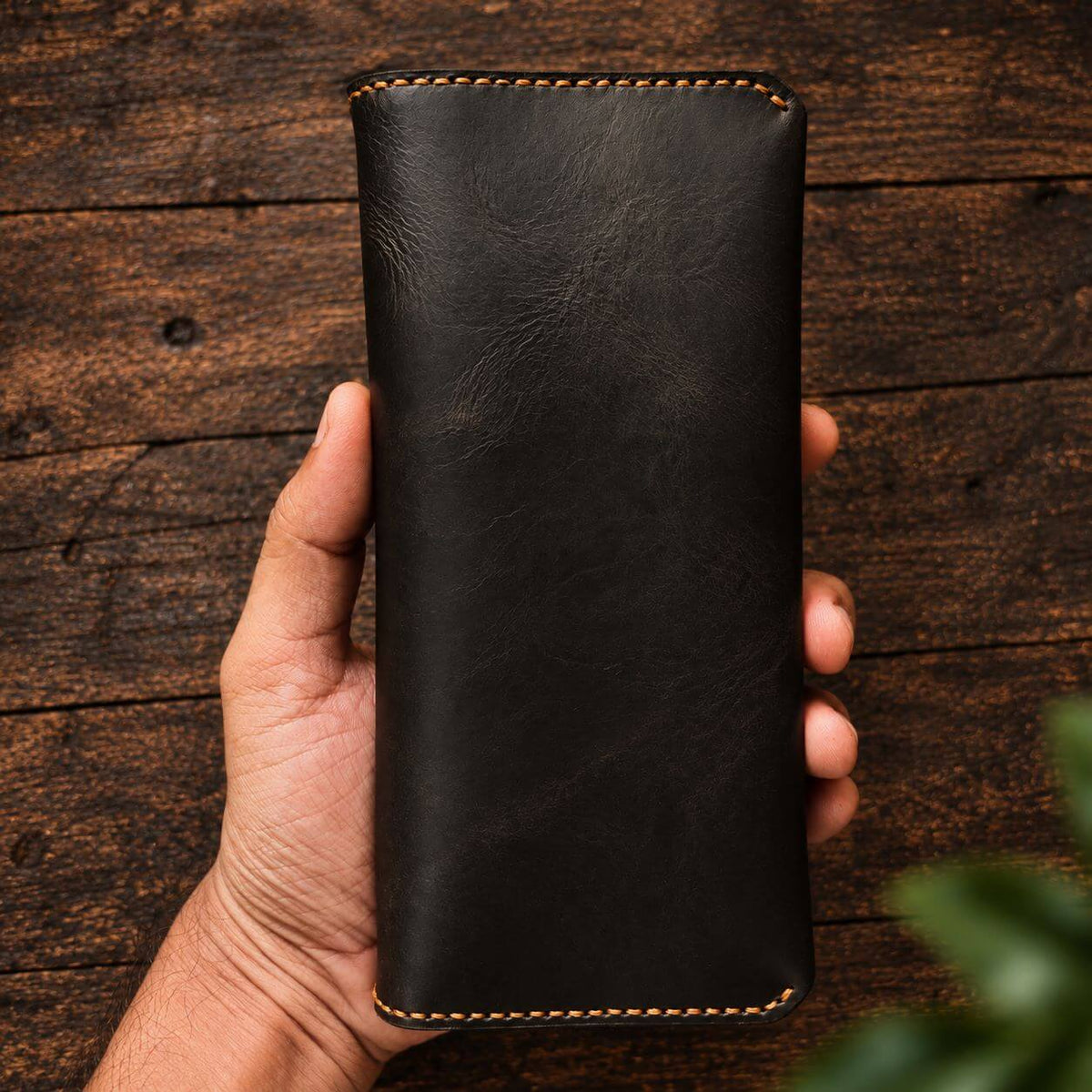 A hand holding a long black leather wallet or card holder with visible tan stitching, set against a rustic dark wood background.