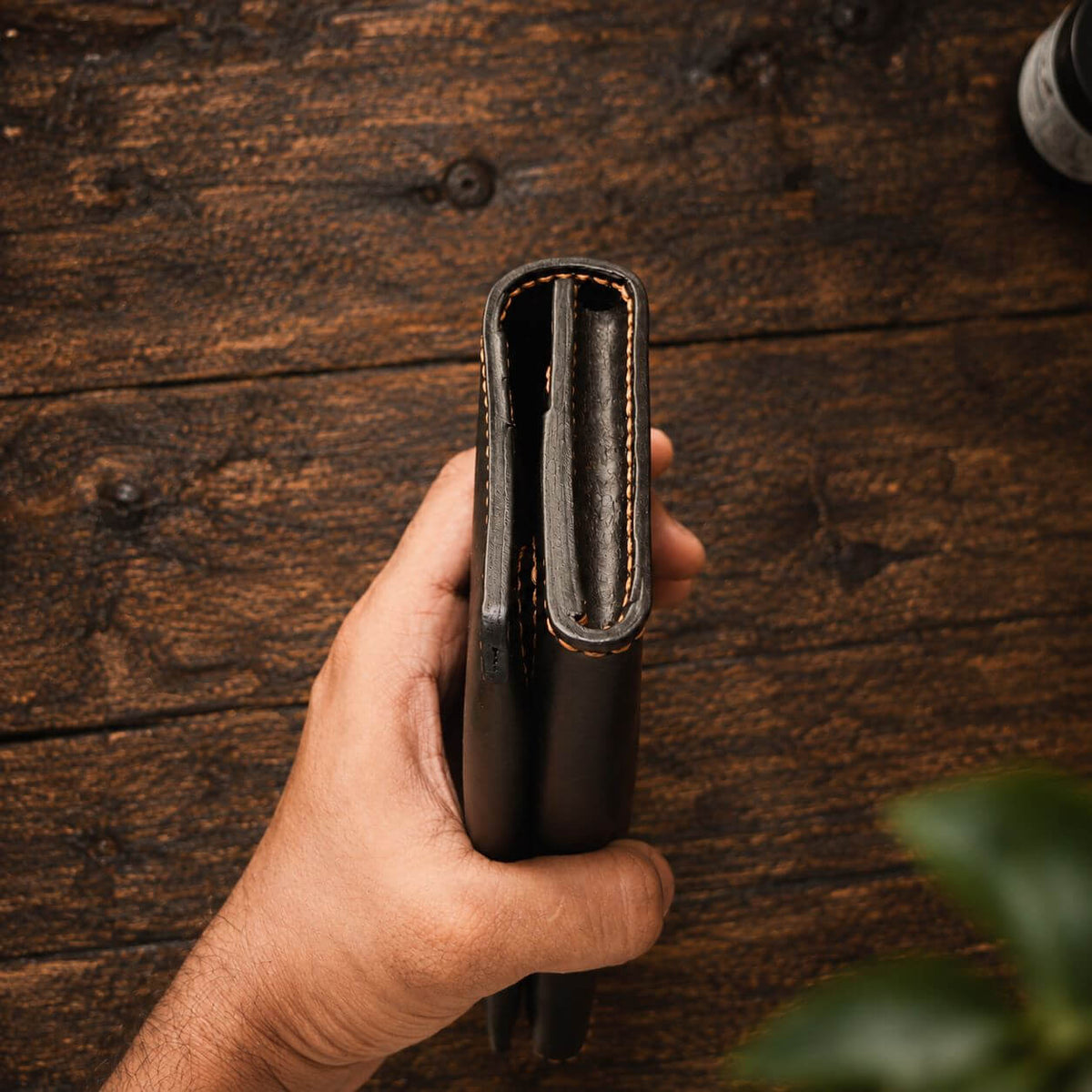 A hand holding the side profile of a dark brown leather wallet or planner organizer, showing the thickness and stitching, set against a rustic wooden background.